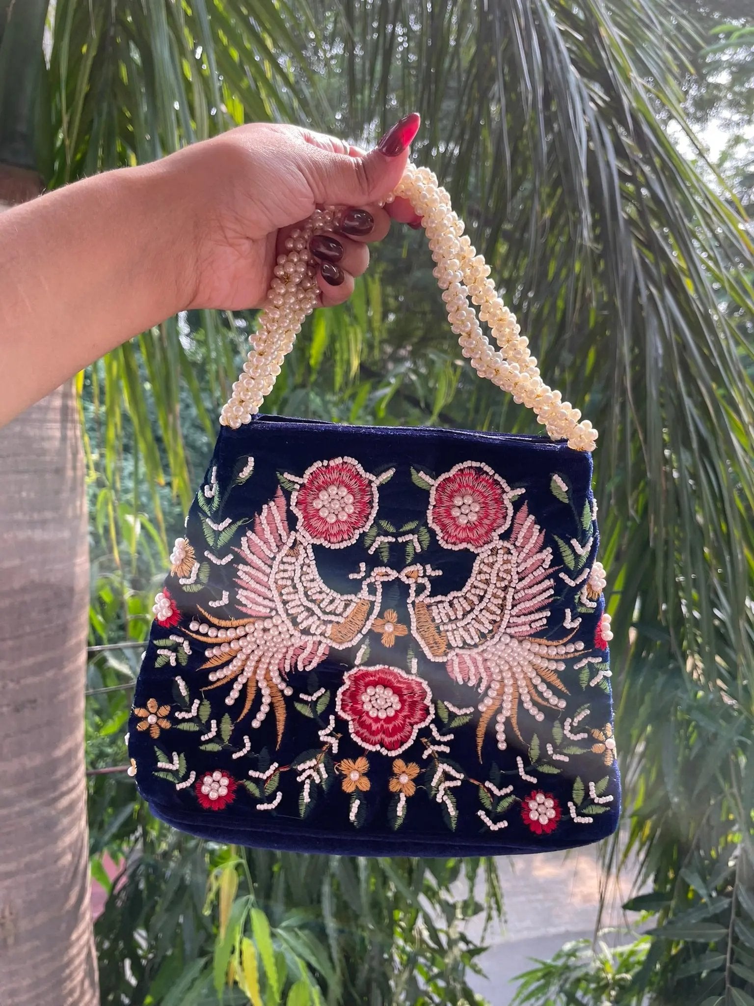 Hand holding a navy blue embroidered handbag with floral and bird designs against a green leafy background.